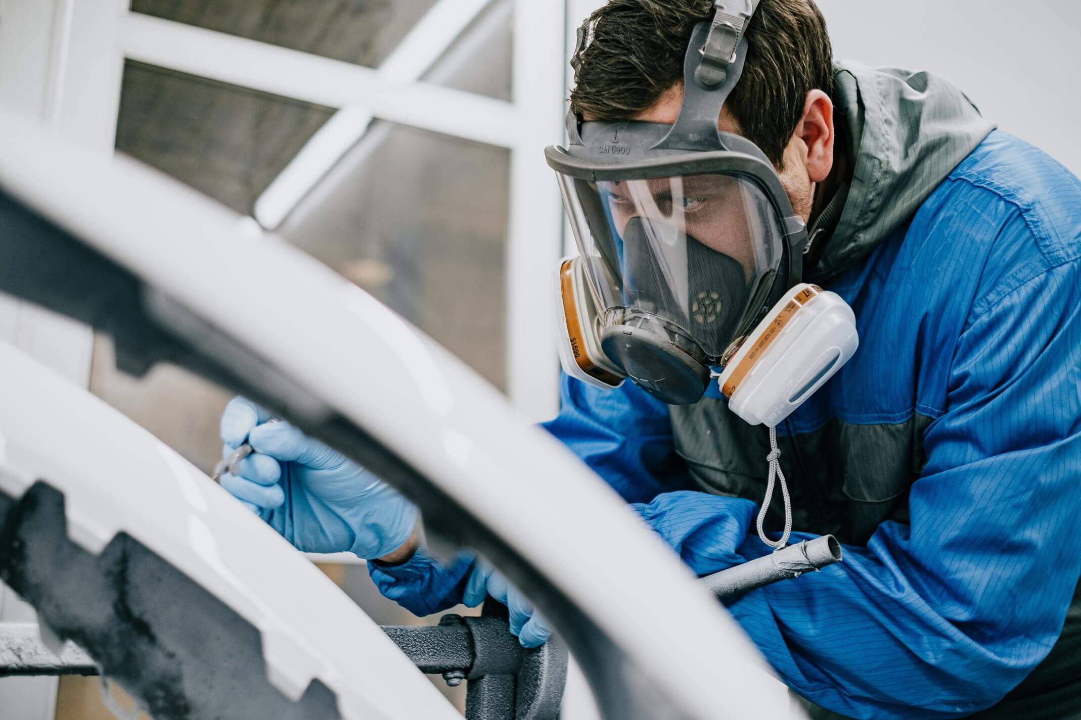 Mitarbeiter mit Schutzmaske und -anzug lackiert in der neuen Lackierhalle der Werkstatt weiße Porscheteile.